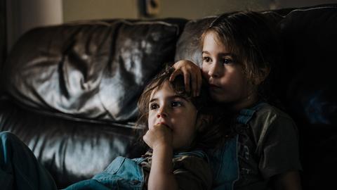 Zwei kleine Mädchen sitzen dicht aneinander gelehnt auf einem Sofa und scheinen fernzusehen.