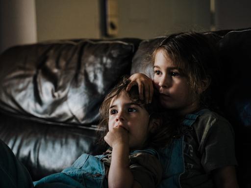 Zwei kleine Mädchen sitzen dicht aneinander gelehnt auf einem Sofa und scheinen fernzusehen.