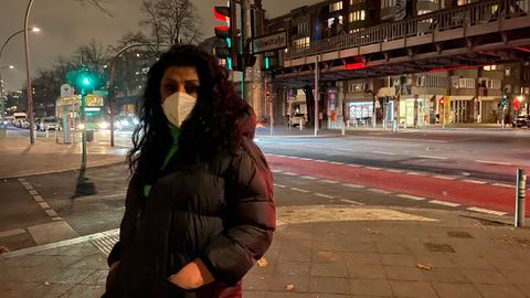 Eine Transsexarbeiterin  mit langen dunklen Haaren und im langen Daunenmantel sowie Mund-Nasenschutz steht an einer Kreuzung in Berlin.