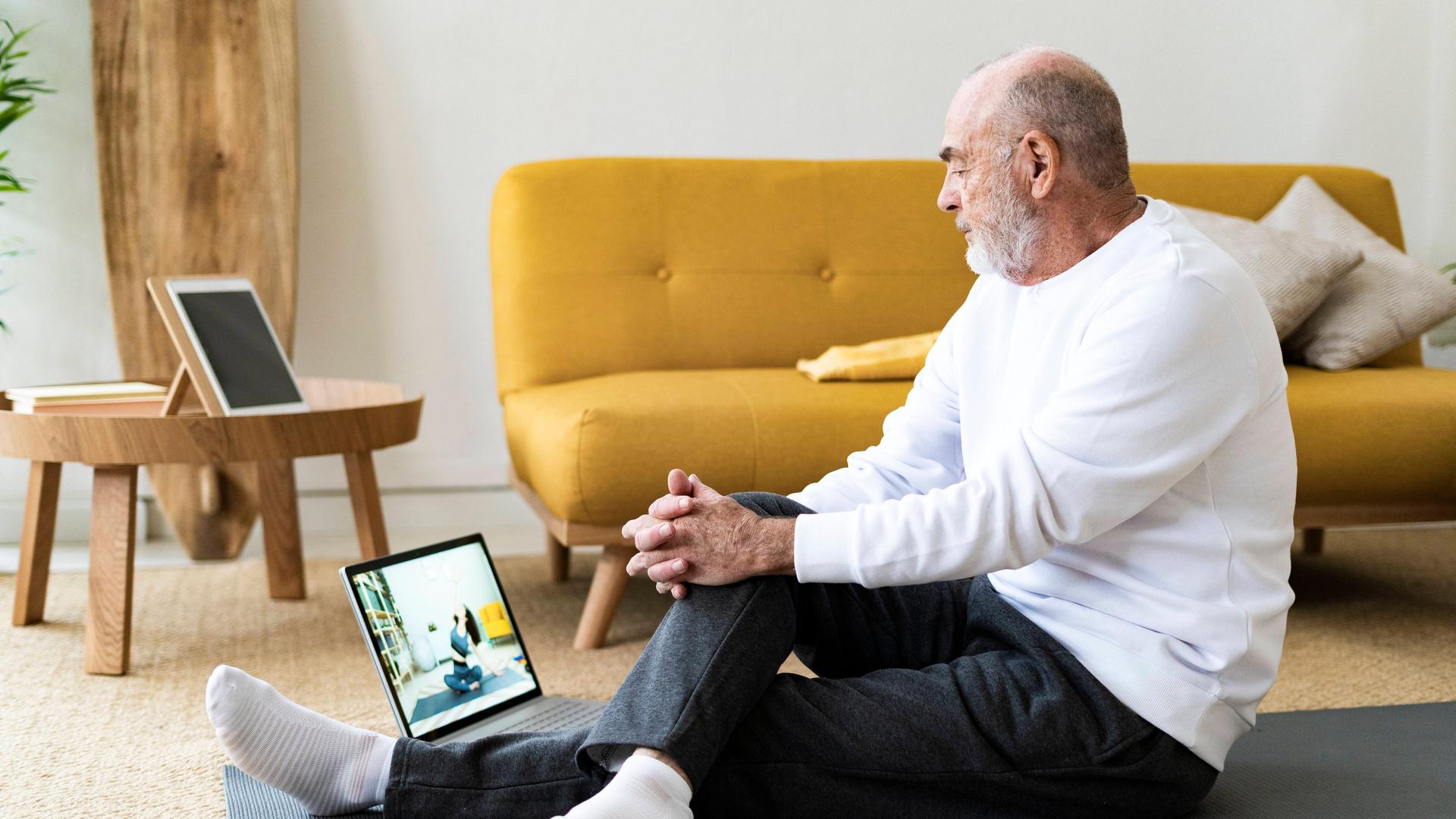 Sport vom Sofa aus: Um fit zu werden, muss man nicht in teure Studios gehen. Findet auch der ältere Mann auf dem Foto, der im bequemen Yoga-Outfit auf einen Laptop-Bildschirm schaut. 