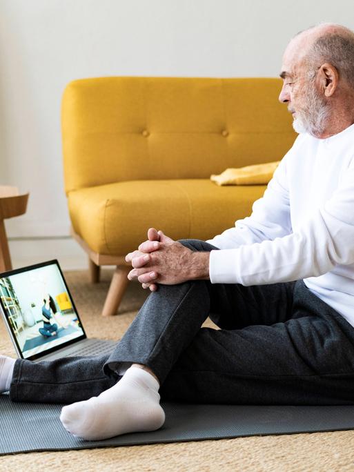 Sport vom Sofa aus: Um fit zu werden, muss man nicht in teure Studios gehen. Findet auch der ältere Mann auf dem Foto, der im bequemen Yoga-Outfit auf einen Laptop-Bildschirm schaut. 