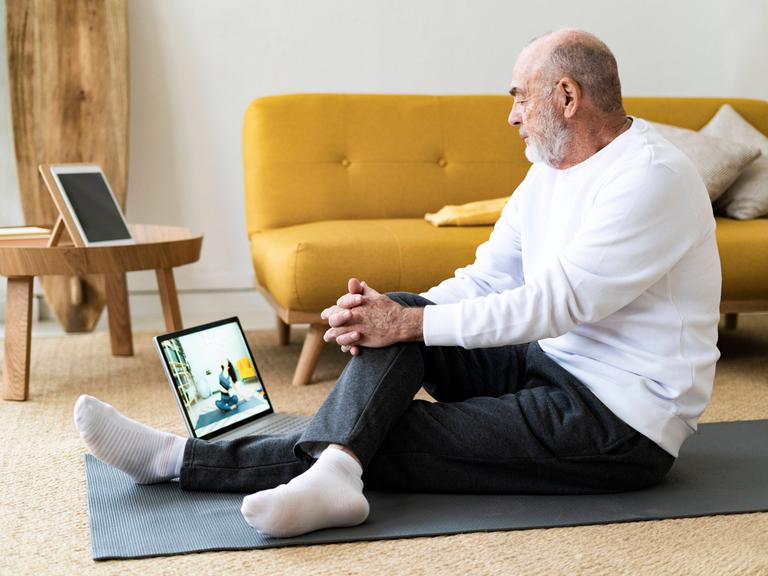 Sport vom Sofa aus: Um fit zu werden, muss man nicht in teure Studios gehen. Findet auch der ältere Mann auf dem Foto, der im bequemen Yoga-Outfit auf einen Laptop-Bildschirm schaut. 