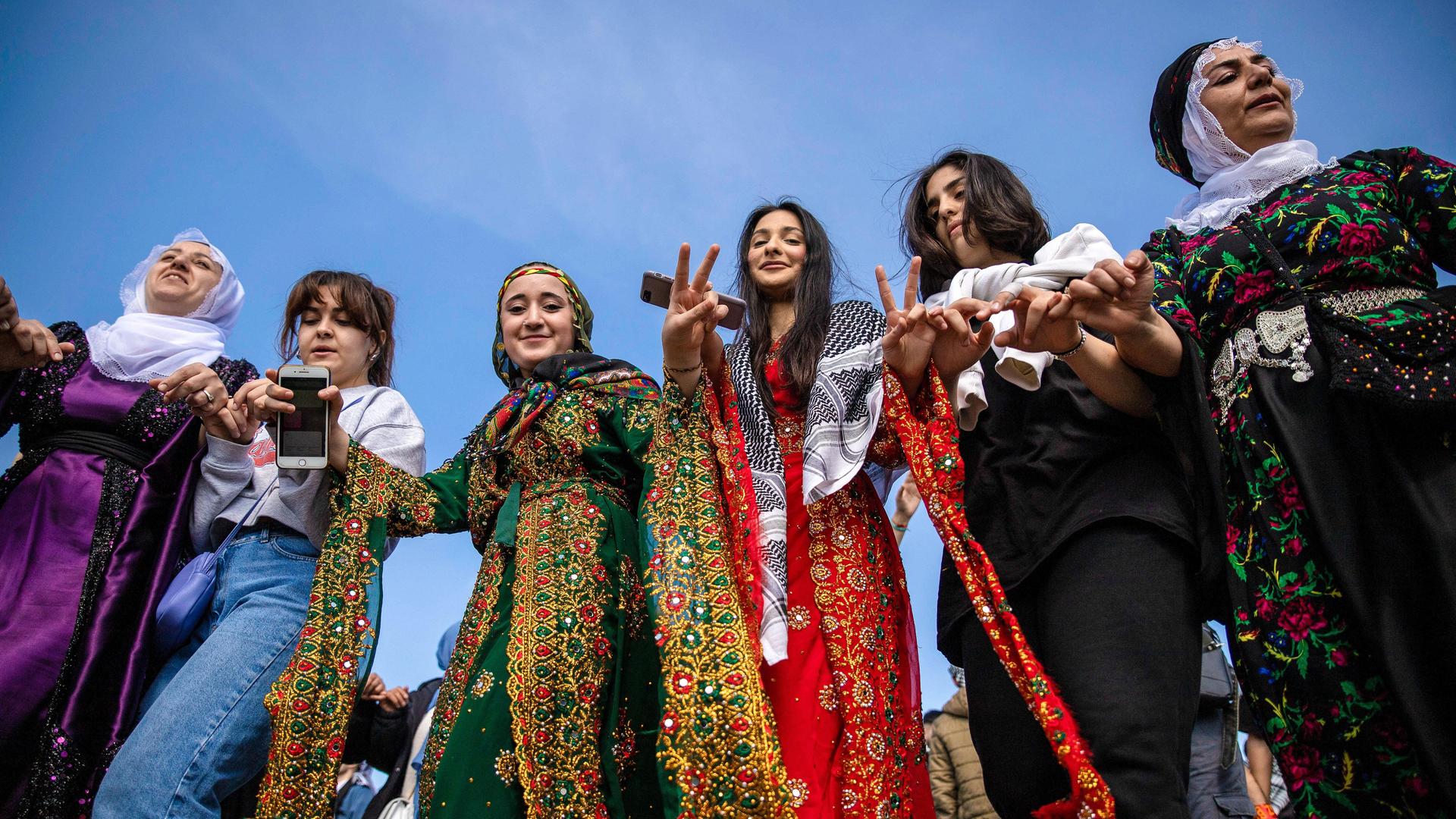 Frauen in farbenfroher traditioneller Kleidung tanzen gemeinsam. Sie schauen lachend in die Kamera.