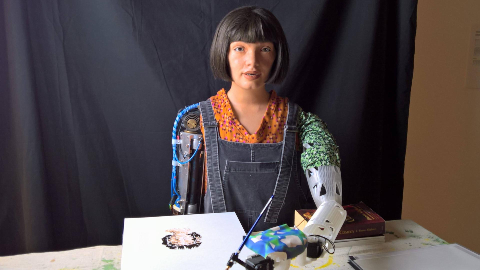 Der humanoide Roboter Ai-Da malt ein Bild. In der Roboterhand hält er einen Pinsel und hat vor sich ein weißes Blatt auf dem ein Kopfporträt zu sehen ist. Der humanoide Roboter Ai-Da malt ein Bild. In der Roboterhand hält er einen Pinsel und hat vor sich ein weißes Blatt auf dem ein Kopfporträt zu sehen ist.