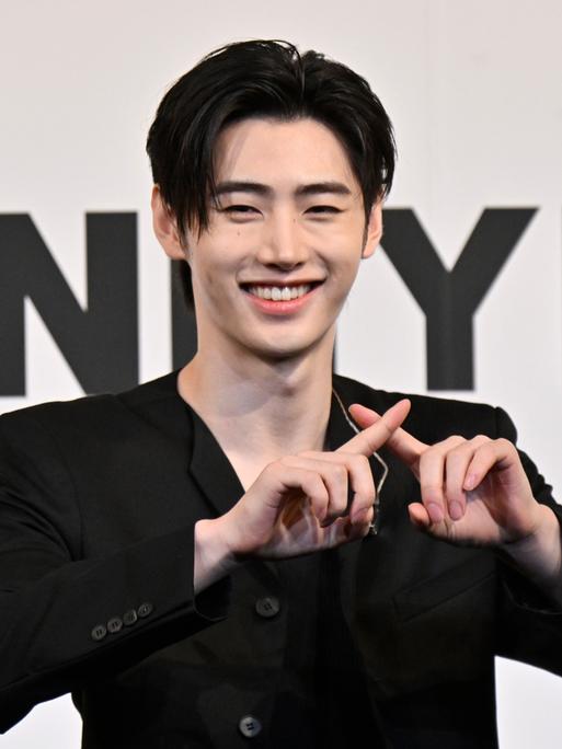 Sänger Sunghoon steht vor dem Logo seiner K-Pop-Gruppe Enhypen, lacht und kreuzt die Finger. 