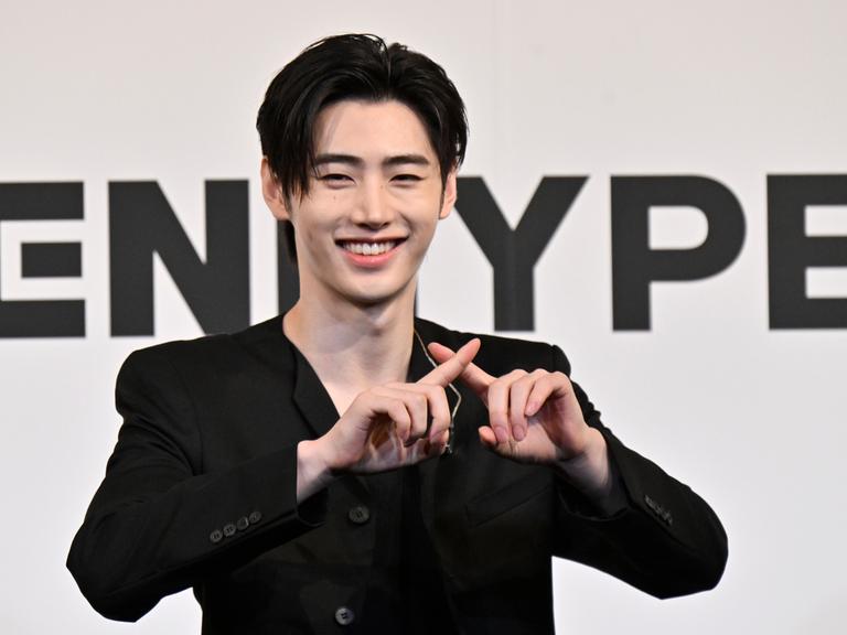 Sänger Sunghoon steht vor dem Logo seiner K-Pop-Gruppe Enhypen, lacht und kreuzt die Finger. 