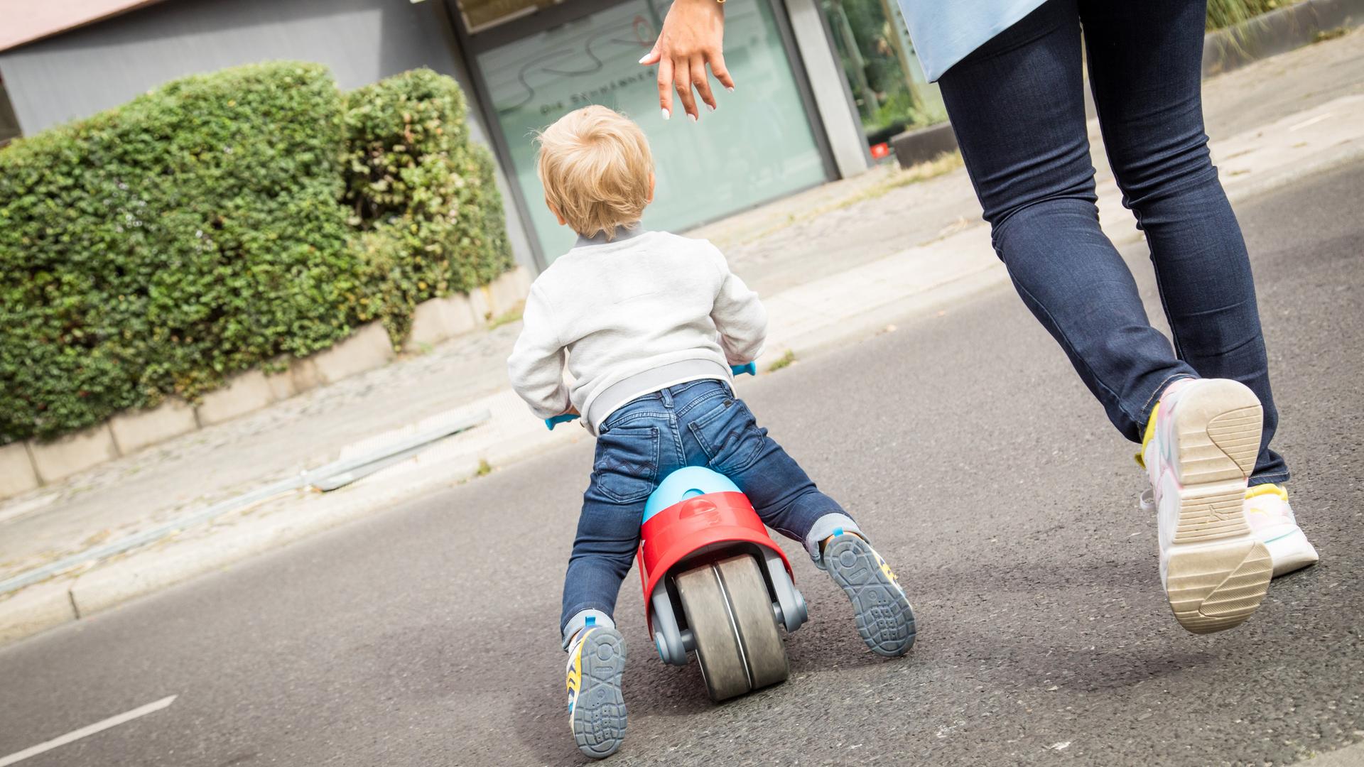 Eine Frau überquert mit einem kleinen Jungen eine Strasse. Er fährt auf einem Lauf-Rad. (gestellte Szene)