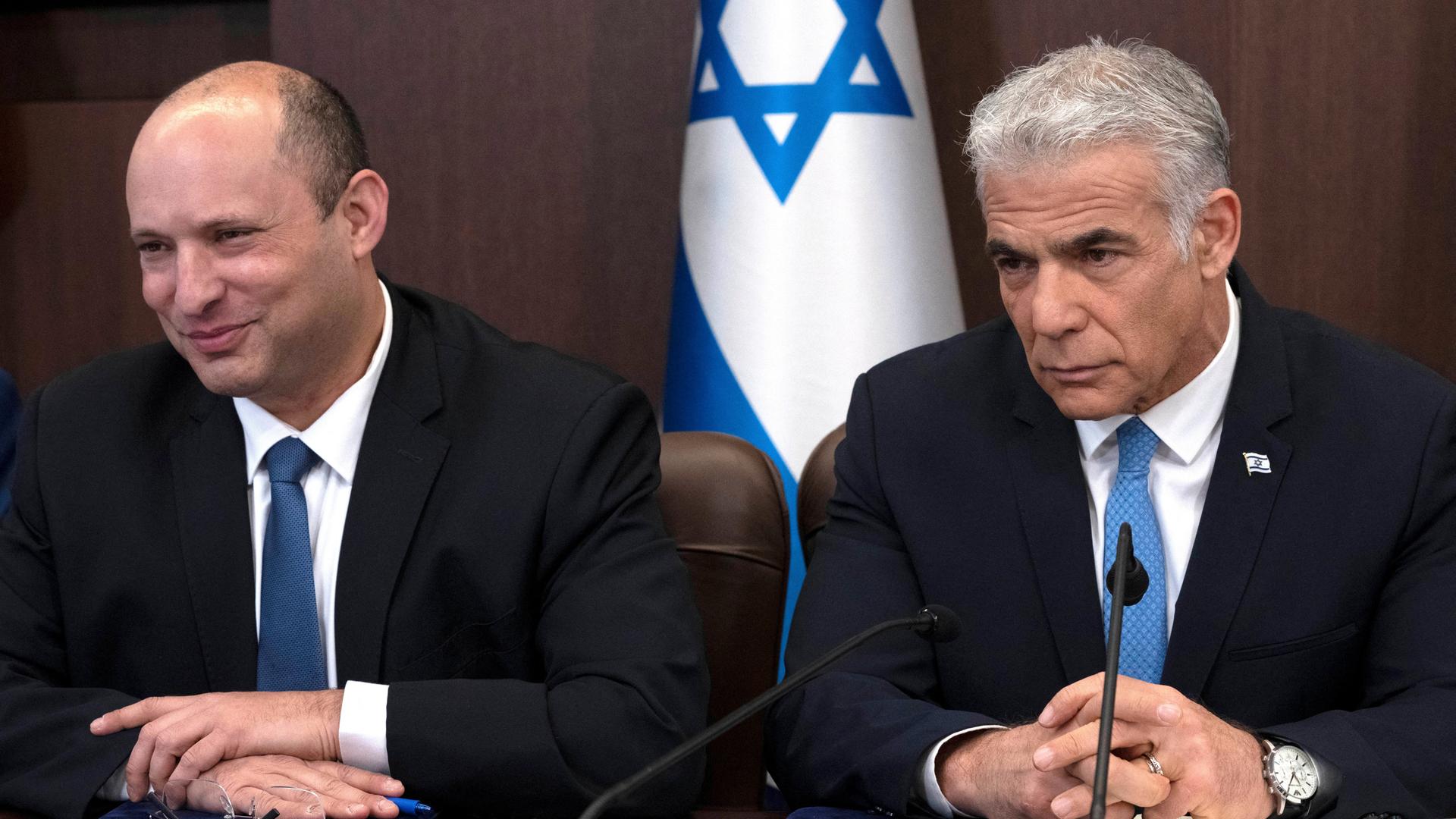 Lapid und Bennett sitzen nebeneinander an einem Tisch. Hinter ihnen steht eine israelische Flagge.