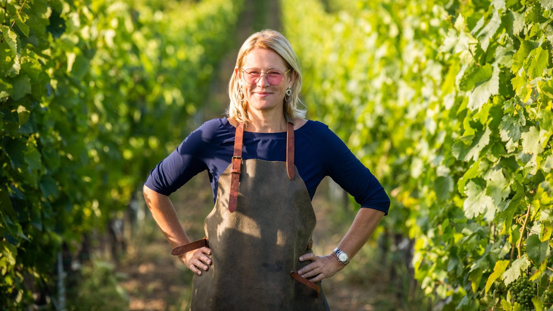 Dorli Muhr steht zwischen Weinreben. Die Hände in die Hüften gestemmt, über der Kleidung eine Schürze. Dorli Muhr steht zwischen Weinreben. Die Hände in die Hüften gestemmt, über der Kleidung eine Schürze.