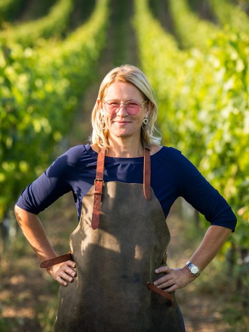 Dorli Muhr steht zwischen Weinreben. Die Hände in die Hüften gestemmt, über der Kleidung eine Schürze.