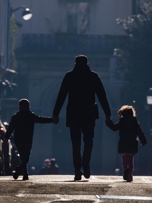 Ein Vater hält seine beiden Kinder an der Hand und geht mit beiden eine Straße entlang.