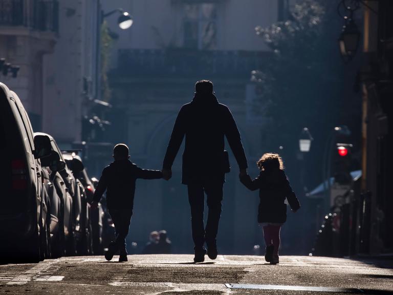 Ein Vater hält seine beiden Kinder an der Hand und geht mit beiden eine Straße entlang.