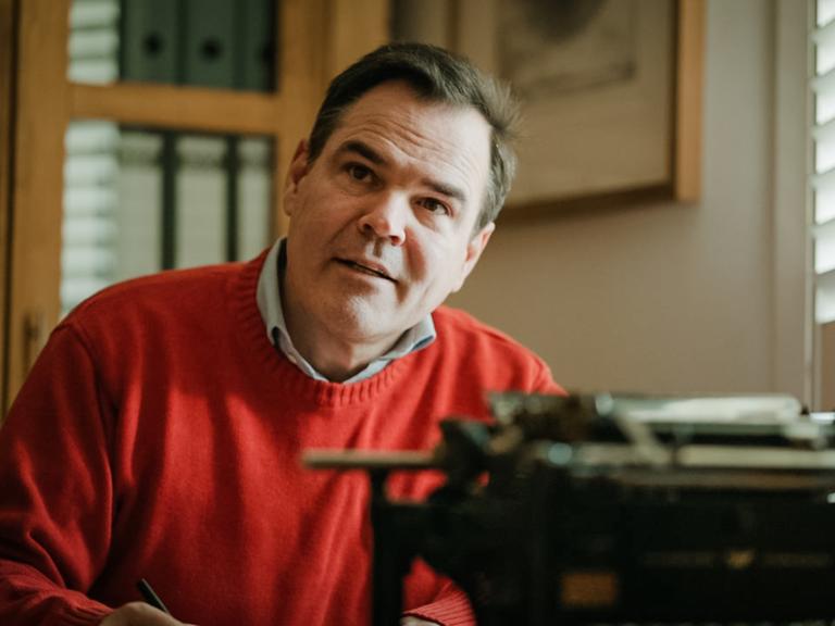 Uwe Tellkamp sitzt im roten Pullover vor einer mechanischen Schreibmaschine und scheint aufmerksam einem Gesprächspartner zuzuhören.