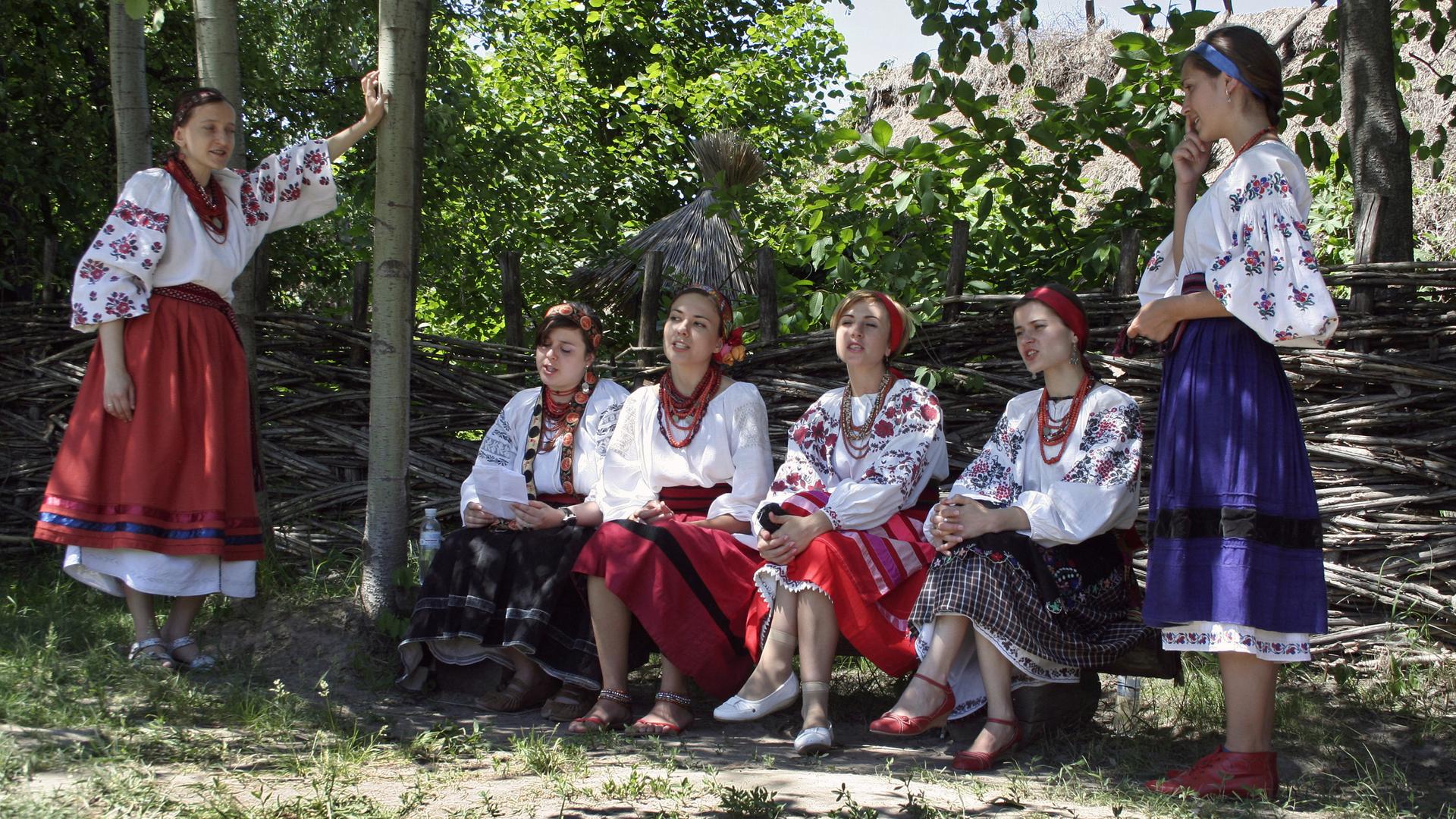 Junge ukrainische Frauen sitzen in traditioneller weißer und bunt gemusterter Kleidung auf einer Bank im Schatten und singen.