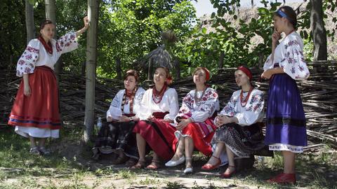 Junge ukrainische Frauen sitzen in traditioneller weißer und bunt gemusterter Kleidung auf einer Bank im Schatten und singen.