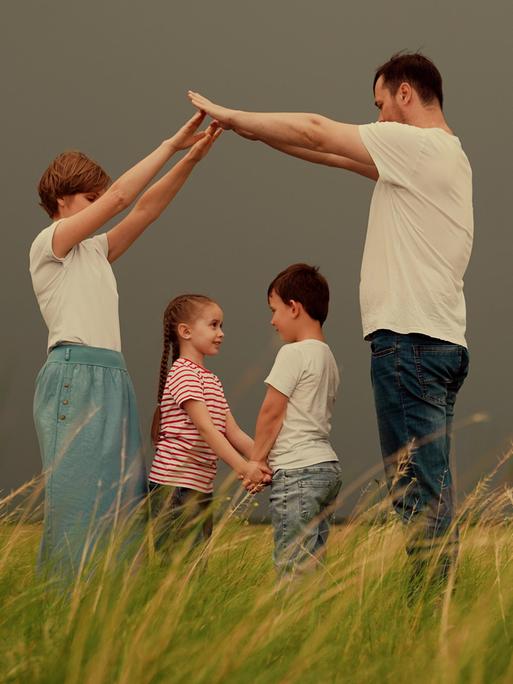 Bild einer Familie mit zwei Kindern, die auf einer Wiese stehen. Mutter und Vater formen mit den Armen ein Dach über dem Kopf der Kinder.