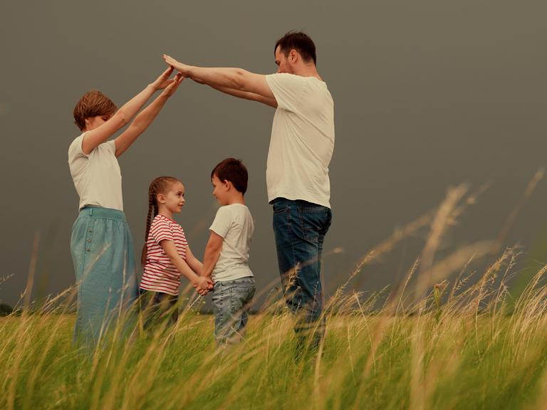 Bild einer Familie mit zwei Kindern, die auf einer Wiese stehen. Mutter und Vater formen mit den Armen ein Dach über dem Kopf der Kinder.