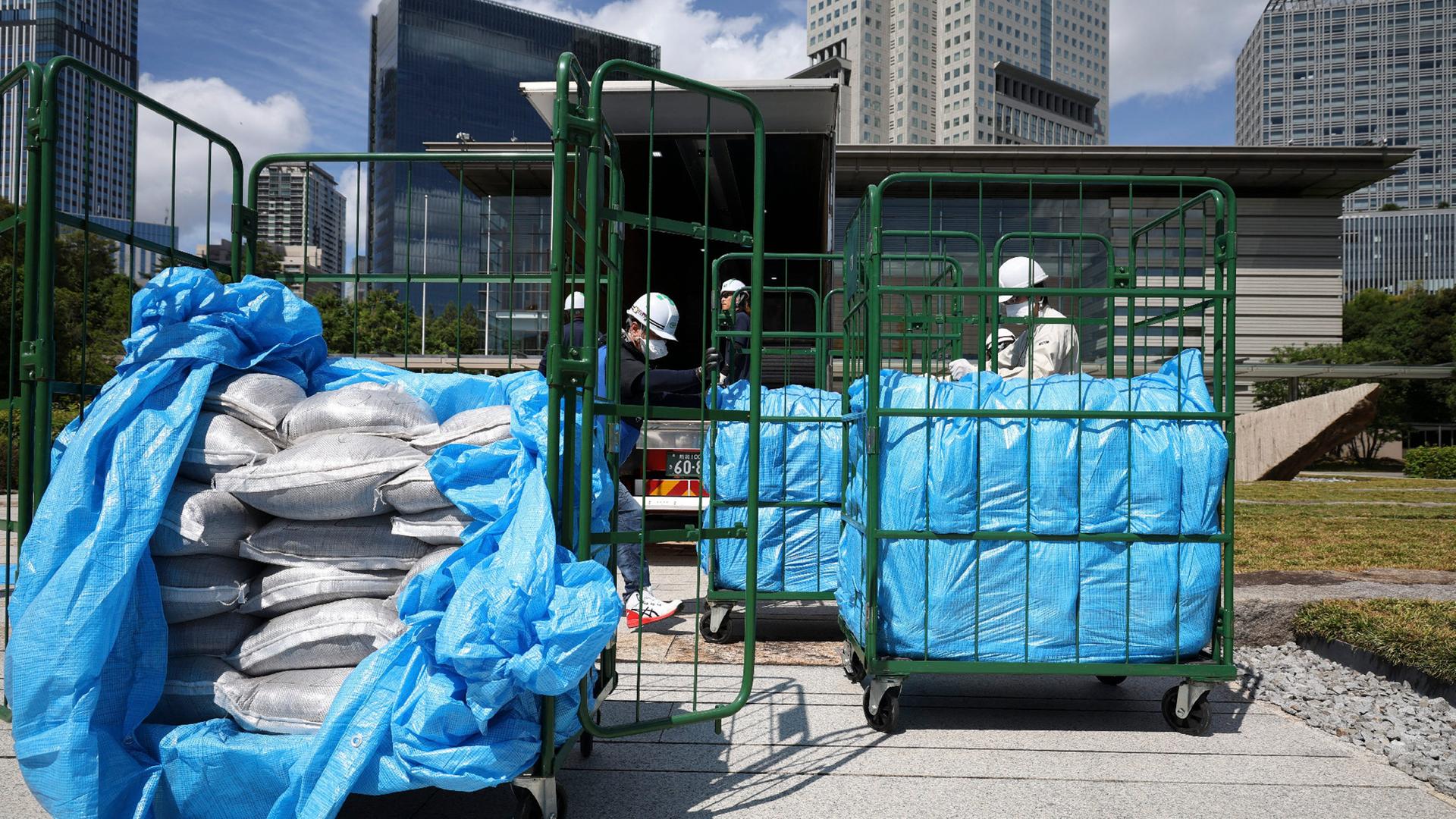 Säcke mit Erde werden in großen Rollcontainern transportiert. Die Arbeiter tragen Schutzkleidung mit Helm und Masken.