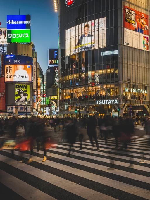 In Tokio überqueren viele Menschen eine Kreuzung, ihre Körper sind Silhouetten.