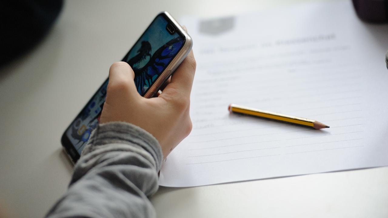 Eine Schülerin hält ein Smartphone in der Hand, Stift und Zettel liegen neben ihr