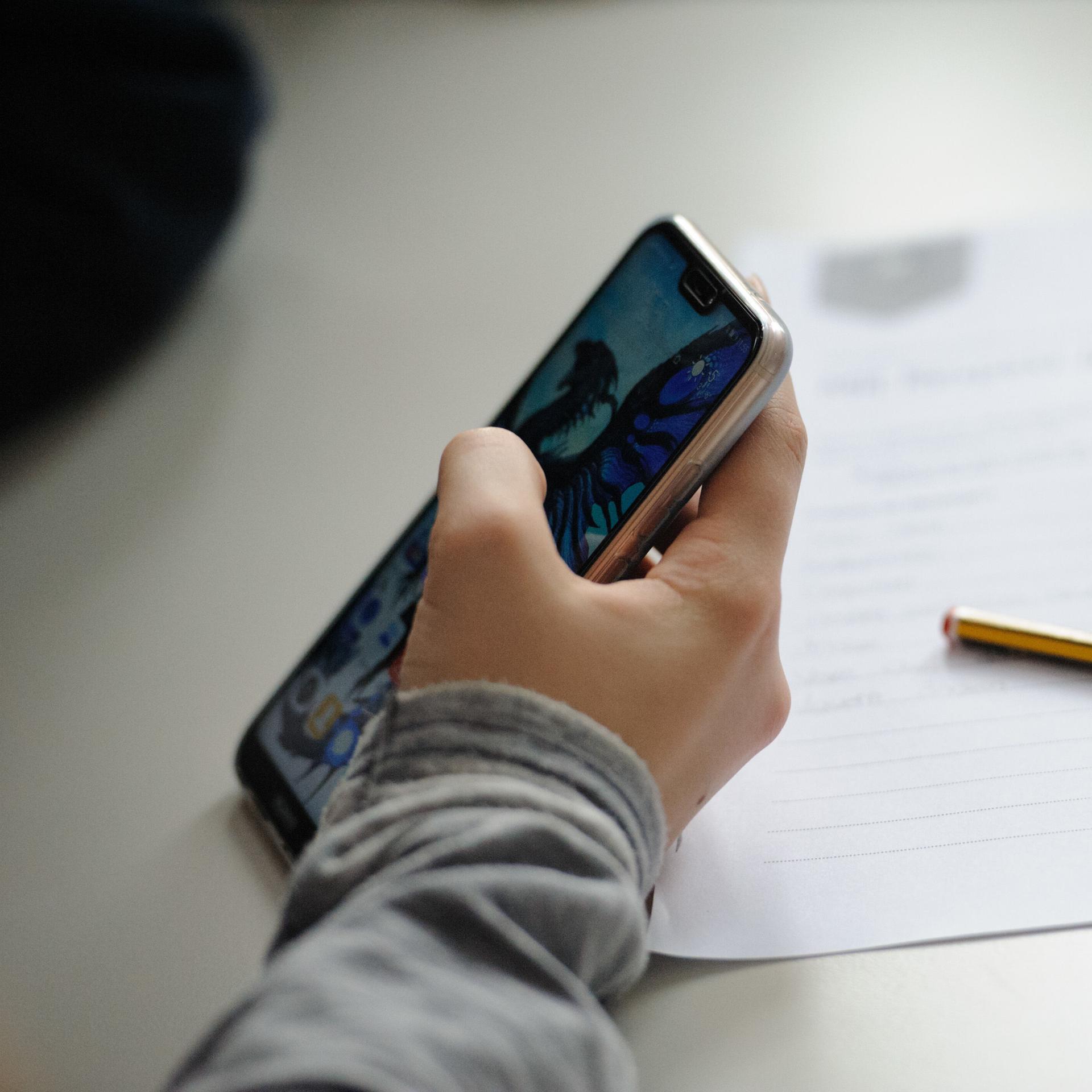 Eine Schülerin hält ein Smartphone in der Hand, Stift und Zettel liegen neben ihr