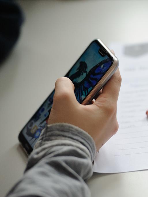 Eine Schülerin hält ein Smartphone in der Hand, Stift und Zettel liegen neben ihr