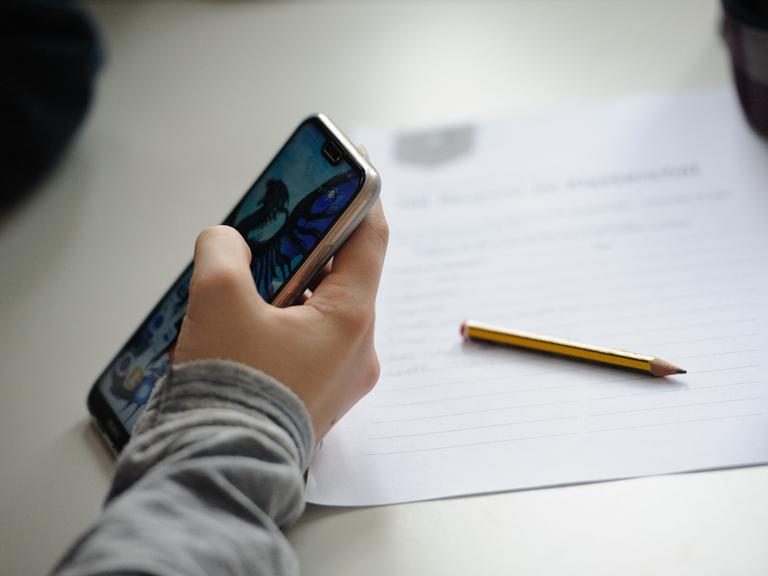 Eine Schülerin hält ein Smartphone in der Hand, Stift und Zettel liegen neben ihr