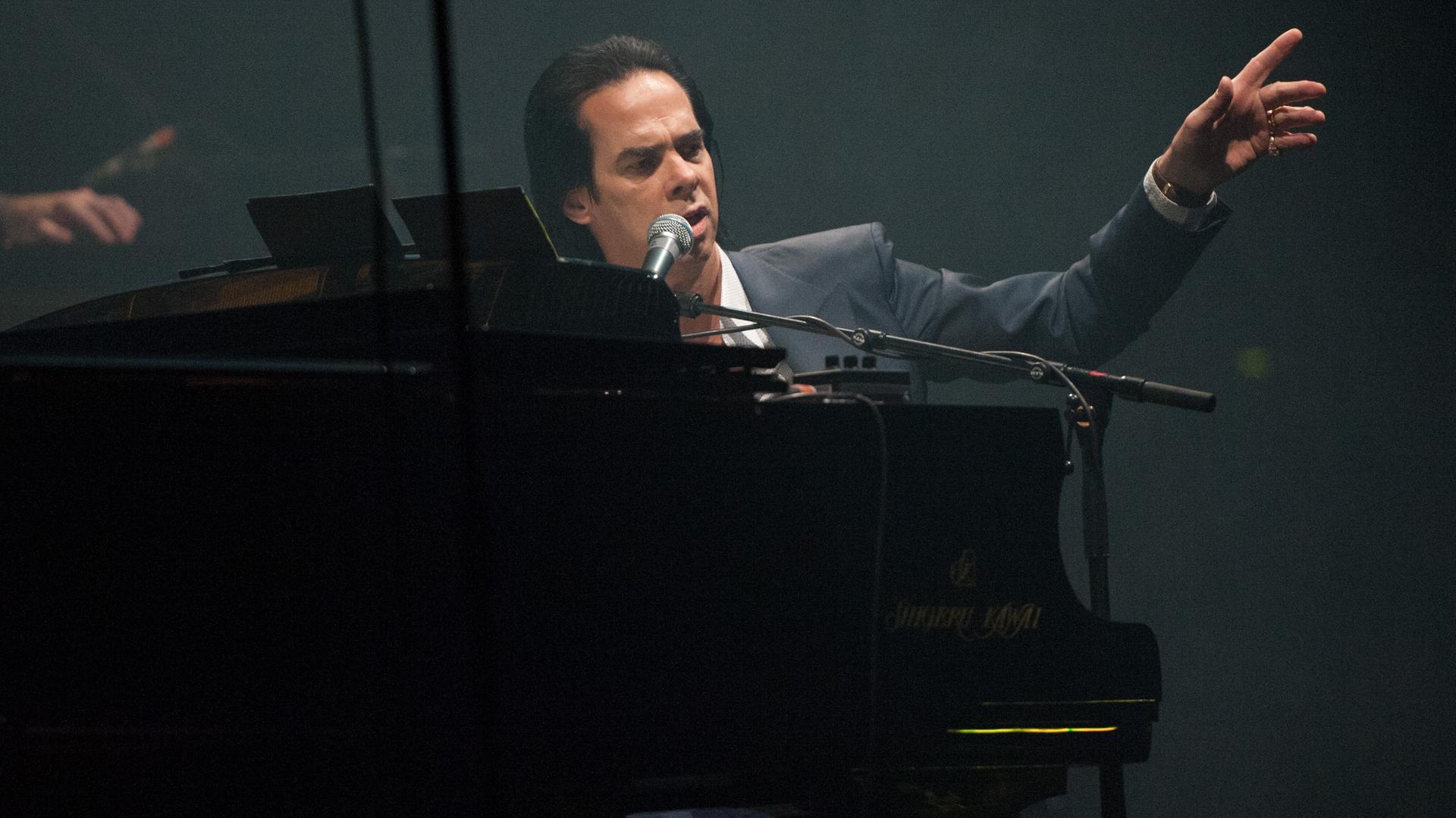 Der australische Sänger Nick Cave sitzt hinter einem Klavier auf einer Bühne und hebt die Hand.