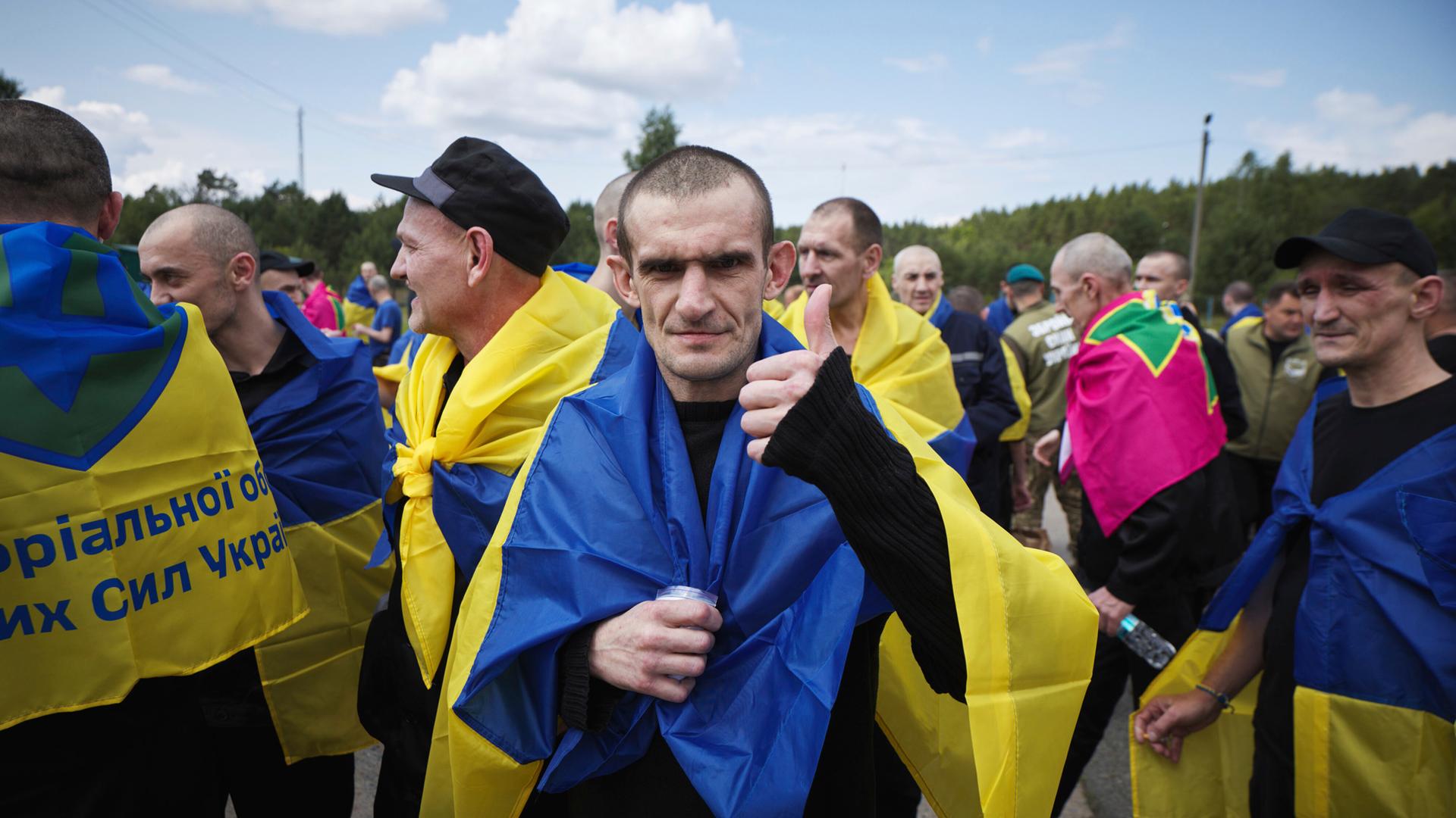 Das Bild zeigt einen ukrainischen Soldaten. Er ist aus russischer Kriegsgefangenschaft zurückgekehrt. Um seine Schultern liegt eine ukrainische Flagge. Er streckt den Daumen hoch. 