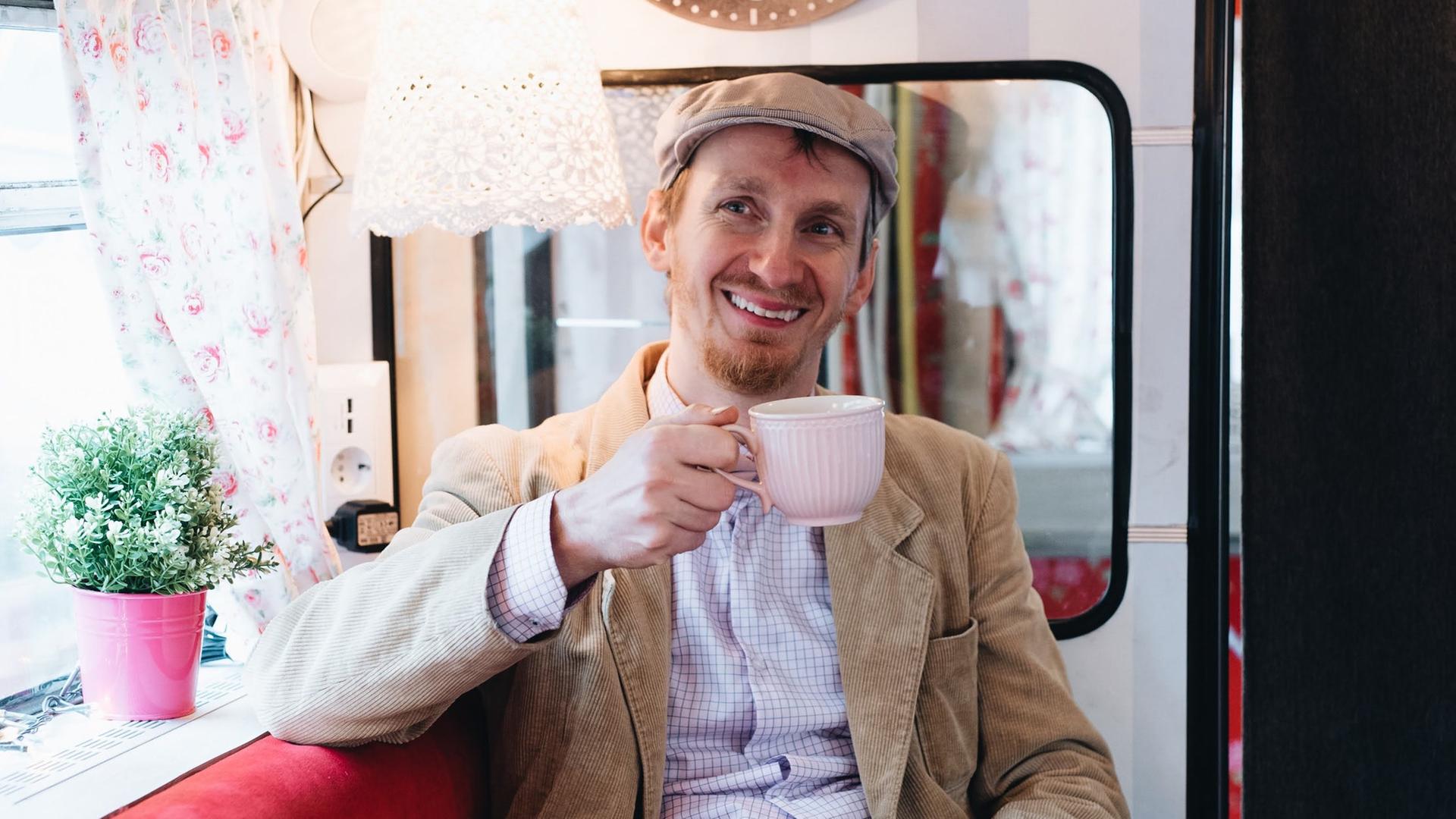 Michèl Malcin sitzt in seinem Bus, hält eine Kaffeetasse in der Hand und lächelt freundlich in die Kamera.