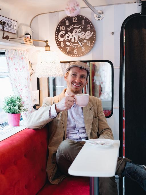 Michèl Malcin sitzt in seinem Bus, hält eine Kaffeetasse in der Hand und lächelt freundlich in die Kamera.