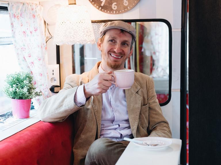 Michèl Malcin sitzt in seinem Bus, hält eine Kaffeetasse in der Hand und lächelt freundlich in die Kamera.