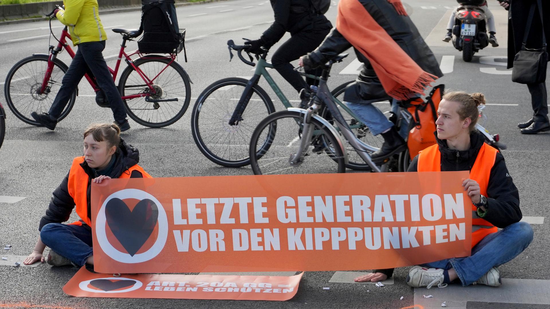 Fahrradfahrer umfahren eine Sitzblockade von zwei Klimaaktivisten. Die beiden halten ein Transparent mit der Aufschrift "Letzte Generation vor den Kipppunkten".