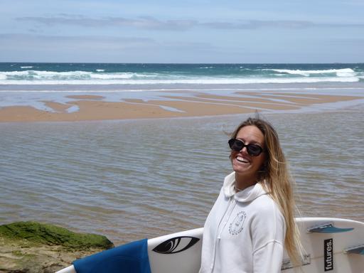 Zu sehen ist Camilla Kemp im Porträt, mit offenen blonden Haaren, einer Sonnenbrille im Gesicht und unterm Arm ihr Surfboard. Im Hintergrund sieht man einen Strand in Portugal.