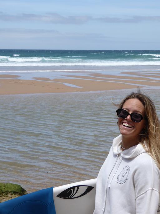 Zu sehen ist Camilla Kemp im Porträt, mit offenen blonden Haaren, einer Sonnenbrille im Gesicht und unterm Arm ihr Surfboard. Im Hintergrund sieht man einen Strand in Portugal.