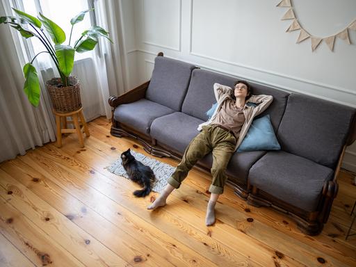 Eine Frau liegt entspannt auf einem grauen Sofa in einem hellen Wohnzimmer. Neben dem Sofa sitzt eine schwarze Katze auf einem Teppich. Links steht eine große grüne Pflanze auf einem Hocker, der Boden ist aus hellem Holz.

