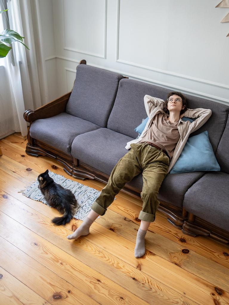 Eine Frau liegt entspannt auf einem grauen Sofa in einem hellen Wohnzimmer. Neben dem Sofa sitzt eine schwarze Katze auf einem Teppich. Links steht eine große grüne Pflanze auf einem Hocker, der Boden ist aus hellem Holz.
