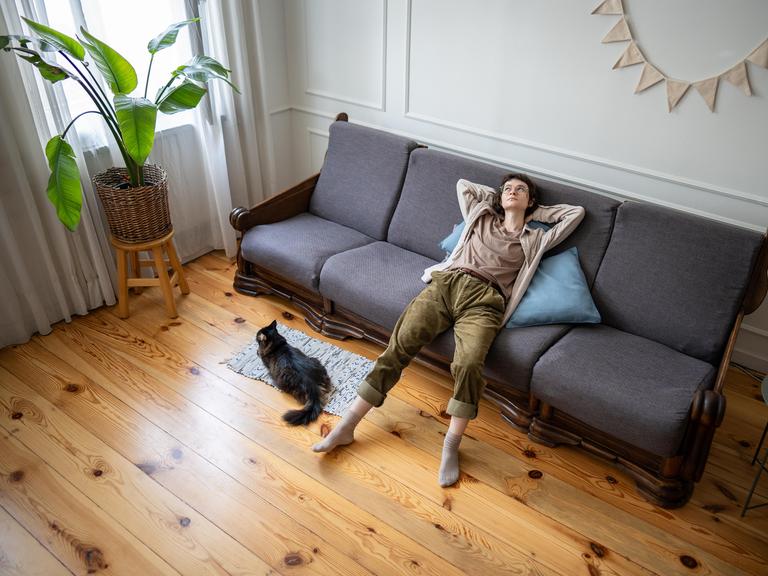 Eine Frau liegt entspannt auf einem grauen Sofa in einem hellen Wohnzimmer. Neben dem Sofa sitzt eine schwarze Katze auf einem Teppich. Links steht eine große grüne Pflanze auf einem Hocker, der Boden ist aus hellem Holz.
