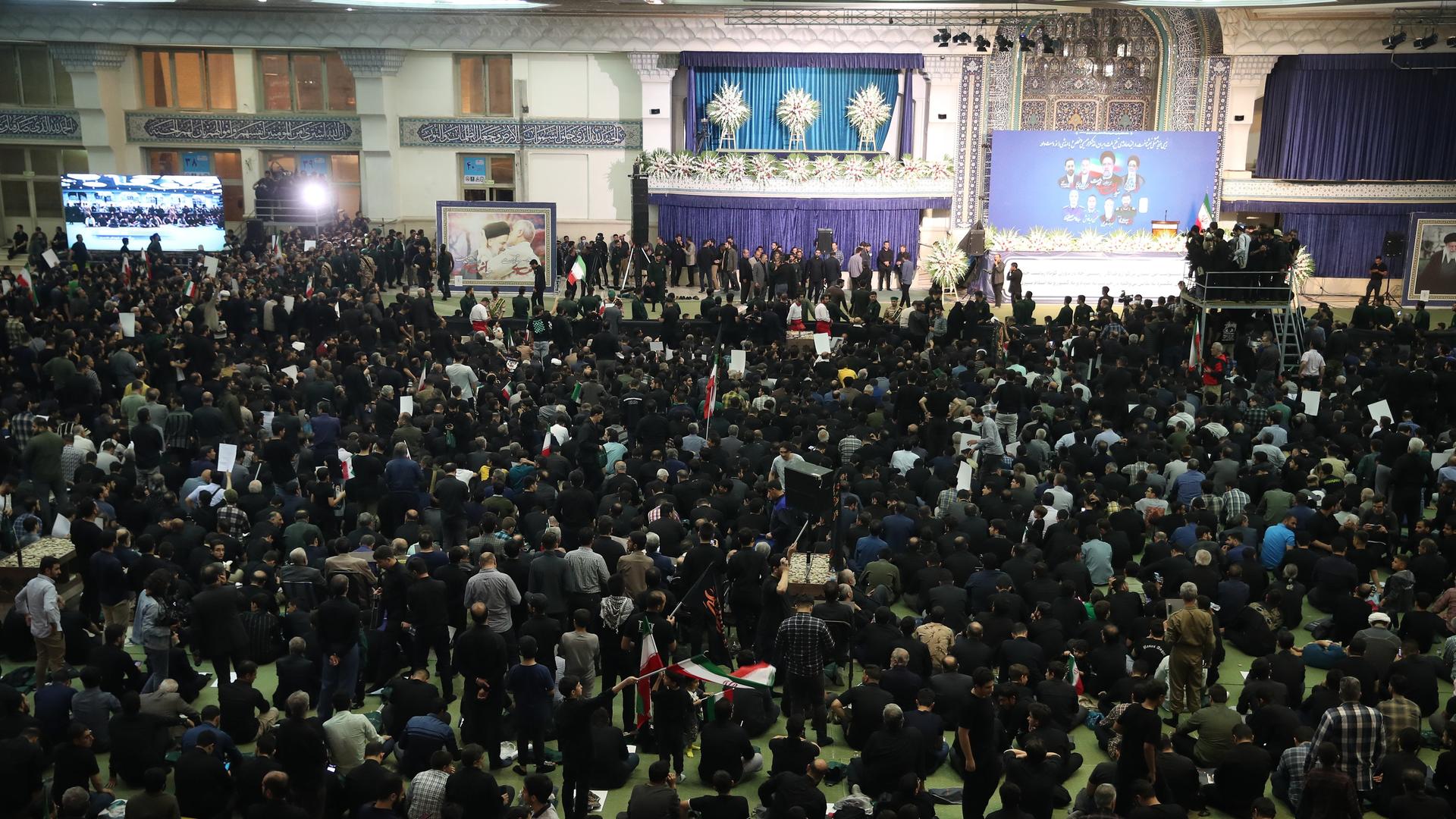 Blick von oben auf hunderte in schwarz gekleidete Menschen in einem großen Raum mit blauen Vorhängen.