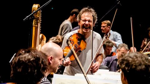 Martial Gauthier hält seine Geige als Linkshänder an die Schulter und lacht dabei in die Kamera im Kreise seiner Musikerkollegen.