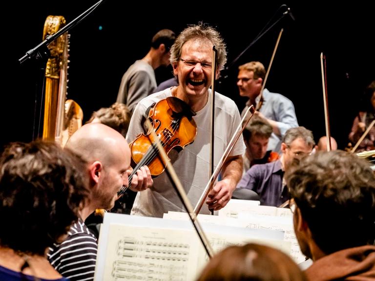 Martial Gauthier hält seine Geige als Linkshänder an die Schulter und lacht dabei in die Kamera im Kreise seiner Musikerkollegen.