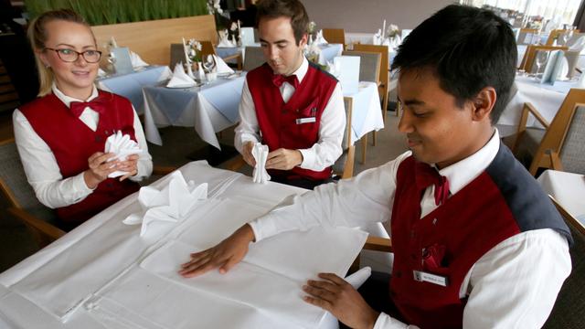 3 Menschen stehen um einen Tisch. Sie falten Servietten. Sie tragen weiße Hemden und rote Westen.