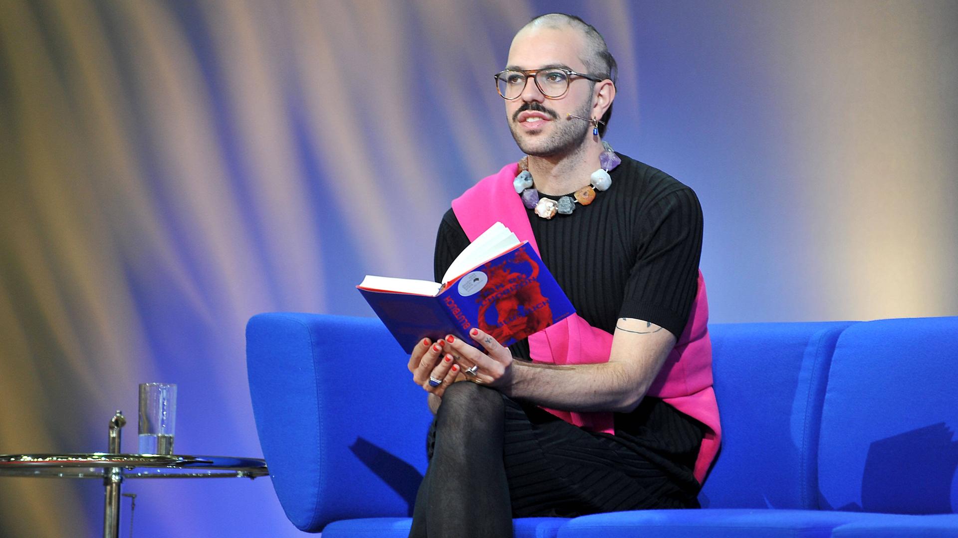 Kim de l'Horizon mit dem Buch "Blutbuch" auf einem knallblauen Sofa sitzend bei der Frankfurter Buchmesse. Kim de l'Horizon mit dem Buch "Blutbuch" auf einem knallblauen Sofa sitzend bei der Frankfurter Buchmesse.