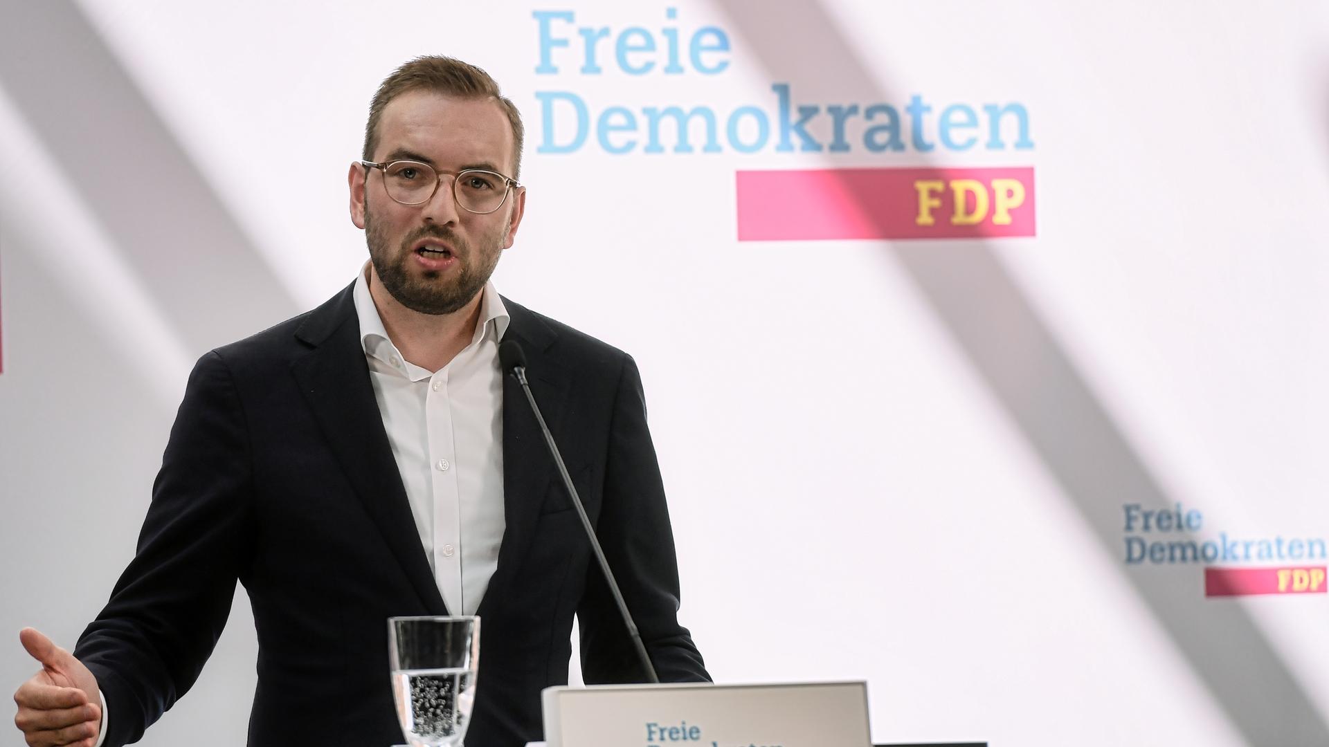 Braun im dunklen Anzug, mit Vollbart und Brille spricht in ein Mikrofon und gestikuliert mit der rechten Hand. Dahinter auf einer weißen Wand der Schriftzug "Freie Demokraten FDP".