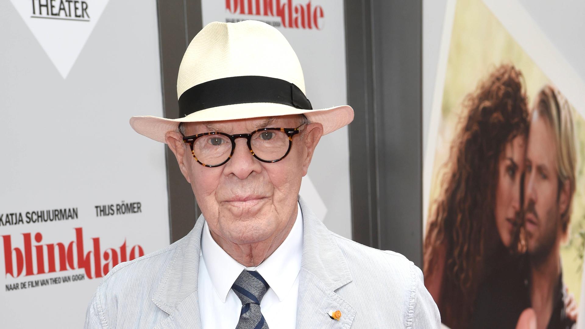 Hans van Manen steht mit weißem Hut vor einem Theatereingang in Amsterdam. 