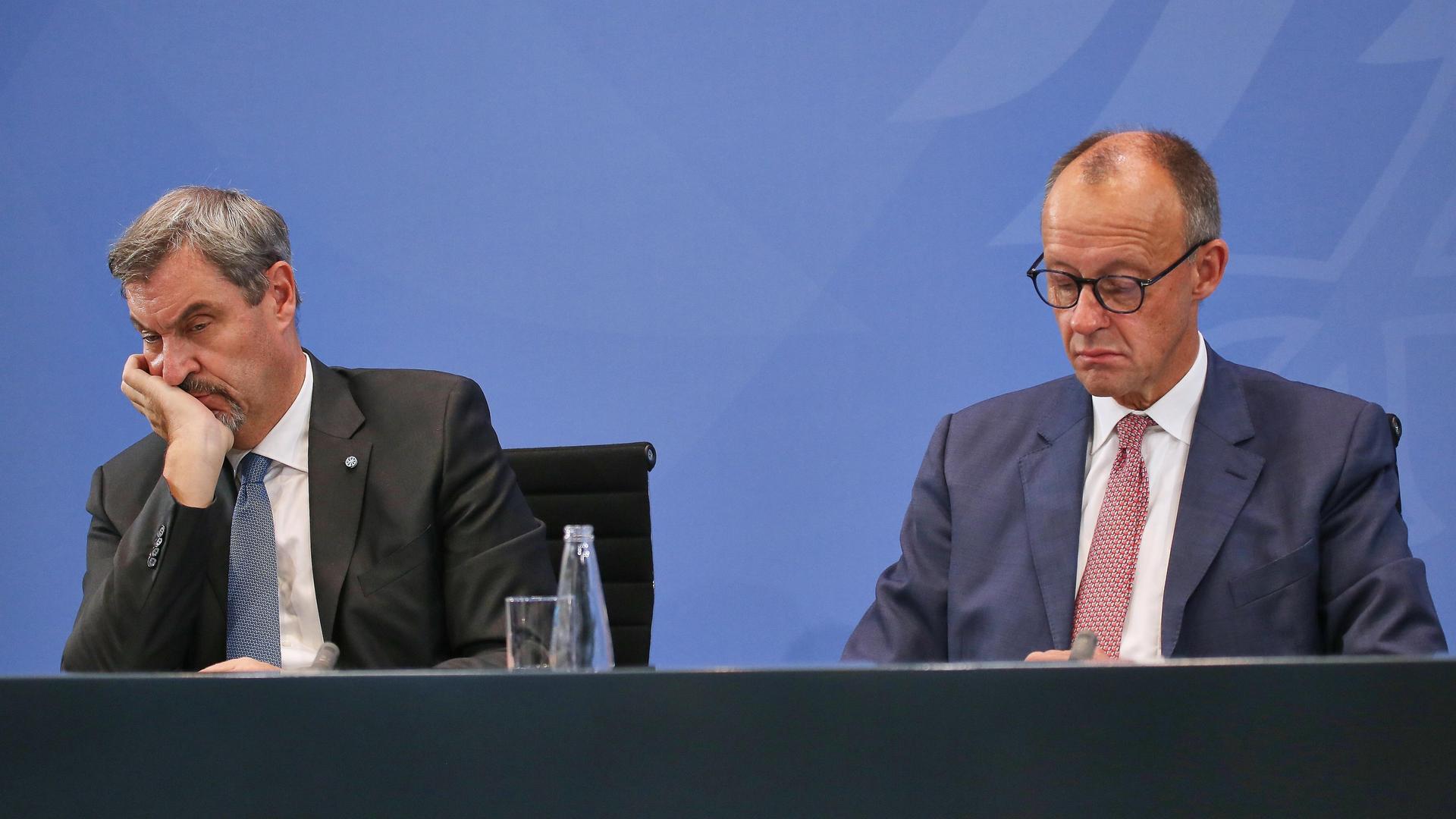 Auf einem Podium sitzen zwei Männer und schauen gelangweilt drein. Der eine trägt einen schwarzen, der andere einen dunkelblauen Anzug. Rechts sitzt Markus Söder von der CSU, links Bundeskanzler Friedrich Merz von der CDU.