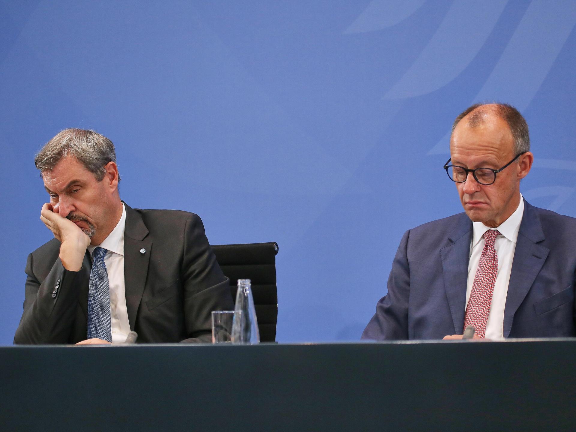 Auf einem Podium sitzen zwei Männer und schauen gelangweilt drein. Der eine trägt einen schwarzen, der andere einen dunkelblauen Anzug. Rechts sitzt Markus Söder von der CSU, links Bundeskanzler Friedrich Merz von der CDU.