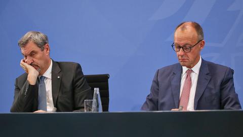 Auf einem Podium sitzen zwei Männer und schauen gelangweilt drein. Der eine trägt einen schwarzen, der andere einen dunkelblauen Anzug. Rechts sitzt Markus Söder von der CSU, links Bundeskanzler Friedrich Merz von der CDU.