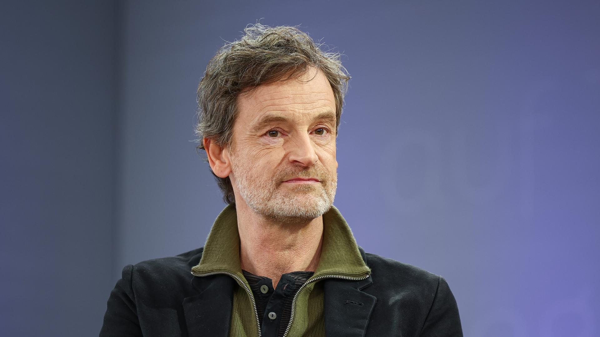 Der deutsche Schauspieler Jörg Hartmann trägt ein schwarzes Sakko und einen grünen Pullover, er sitzt auf einer Bühne mit violettem Hintergrund und schaut freundlich an der Kamera vorbei.