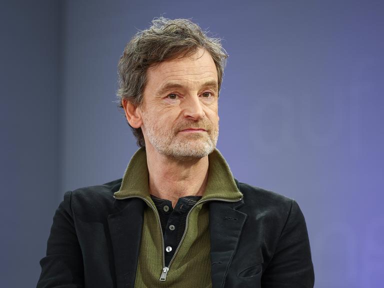 Der deutsche Schauspieler Jörg Hartmann trägt ein schwarzes Sakko und einen grünen Pullover, er sitzt auf einer Bühne mit violettem Hintergrund und schaut freundlich an der Kamera vorbei.